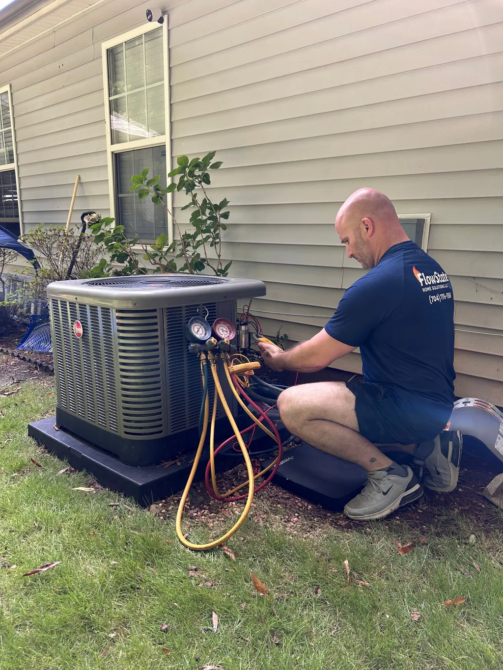 Technician in a branded shirt reading manifold gauges connected to an outdoor condenser unit