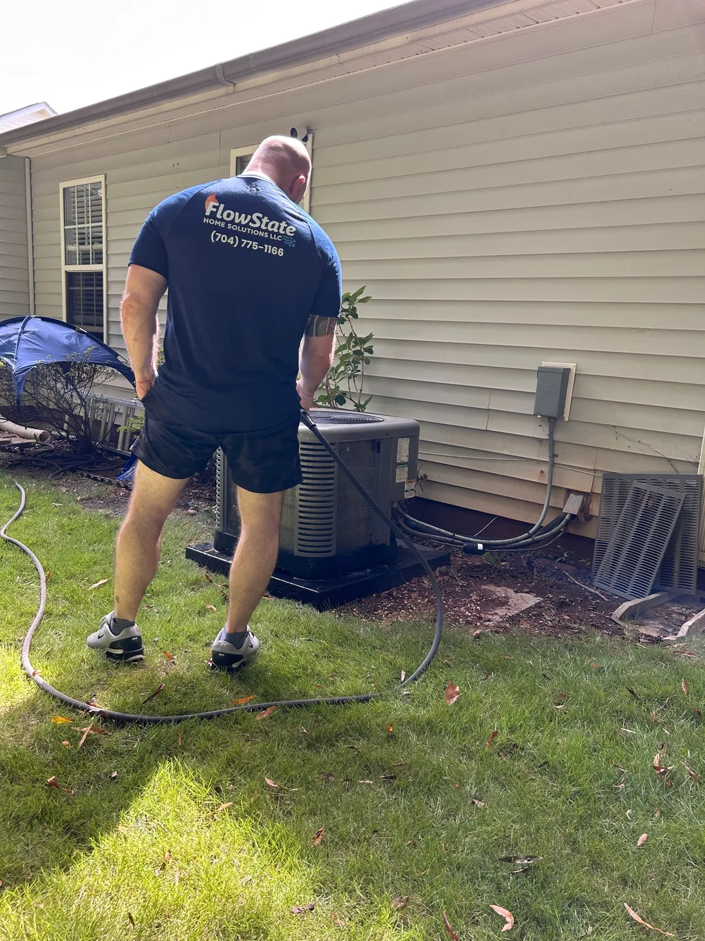 Technician in a FlowState Home Solutions shirt rinsing an outdoor air conditioning condenser beside a house