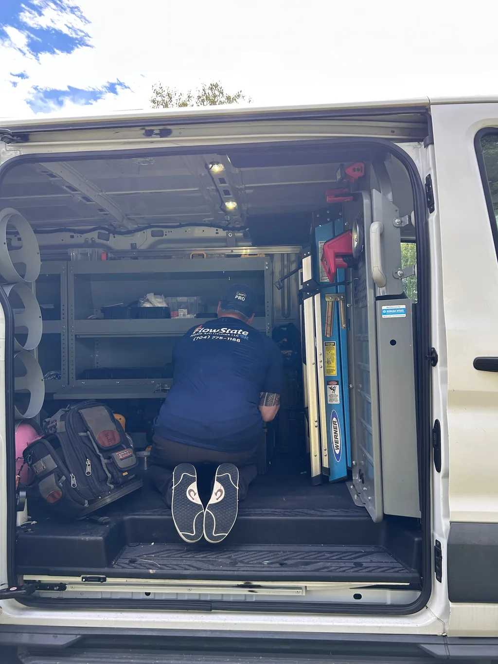 Open work van with parts shelving, a folding ladder and a tool bag, technician kneeling inside